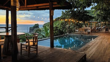 A wooden deck with a pool, lounge chairs, and a view of the ocean at sunset.