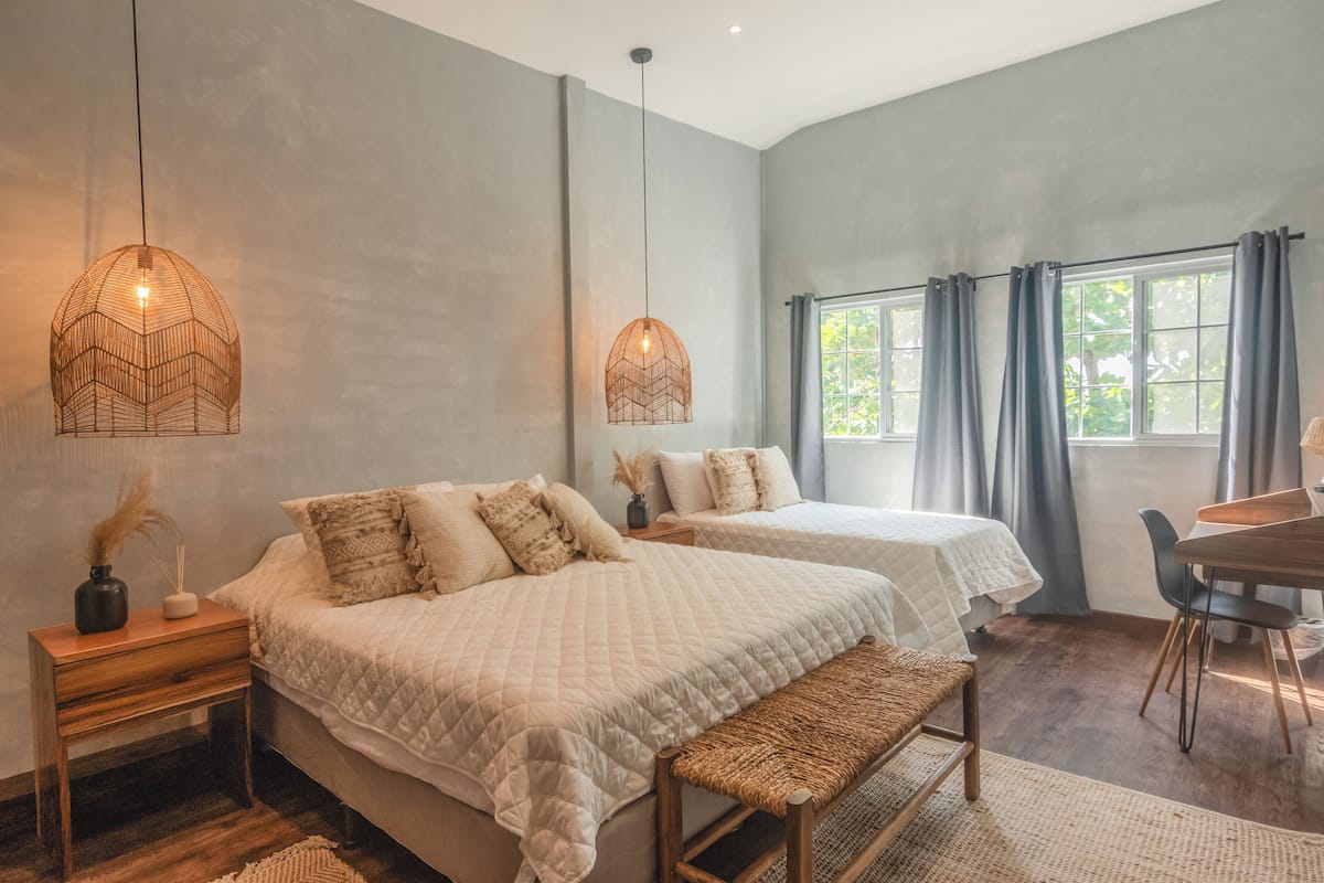 Un dormitorio con dos camas, un banco de madera, una mesita pequeña y una ventana con cortinas.