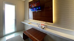 A modern hotel room with a large flat-screen TV mounted on a wooden wall, a small desk with a chair, and a window with a view. - Shero Transpark Bekasi (Bekasi)