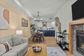 Ein Wohnzimmer mit Sofa, Couchtisch und Kamin. Es gibt eine Essecke mit Tisch und Stühlen sowie eine Küche im Hintergrund.