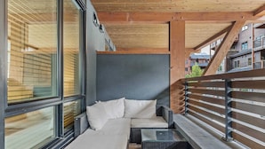 A modern outdoor seating area with a sofa and cushions, a glass table, and a view of the cityscape. - Stylishly Renovated Spring Creek Retreat (Canmore)