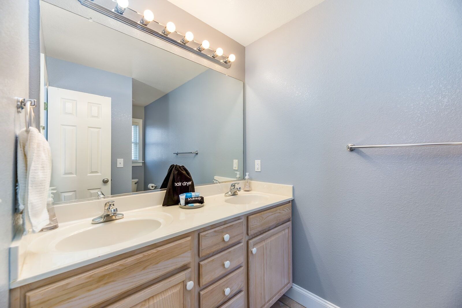 A bathroom with two sinks, a large mirror with lights, a towel rack, and a hair dryer on the counter.