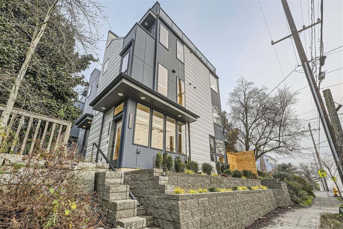 A modern two-story house with a stone staircase leading to the entrance, surrounded by a stone wall and small plants.