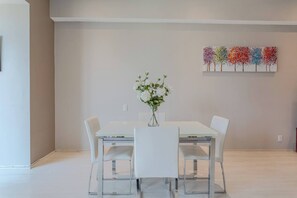 A dining area with a white table and chairs, a vase with flowers, and a colorful tree painting on the wall.