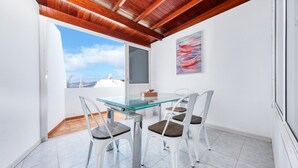 A dining area with a glass table and white chairs, a painting on the wall, and a view of the sky and mountains through a window. - Casa el Varadero - Pool and Gym By LVH (Puerto del Carmen)