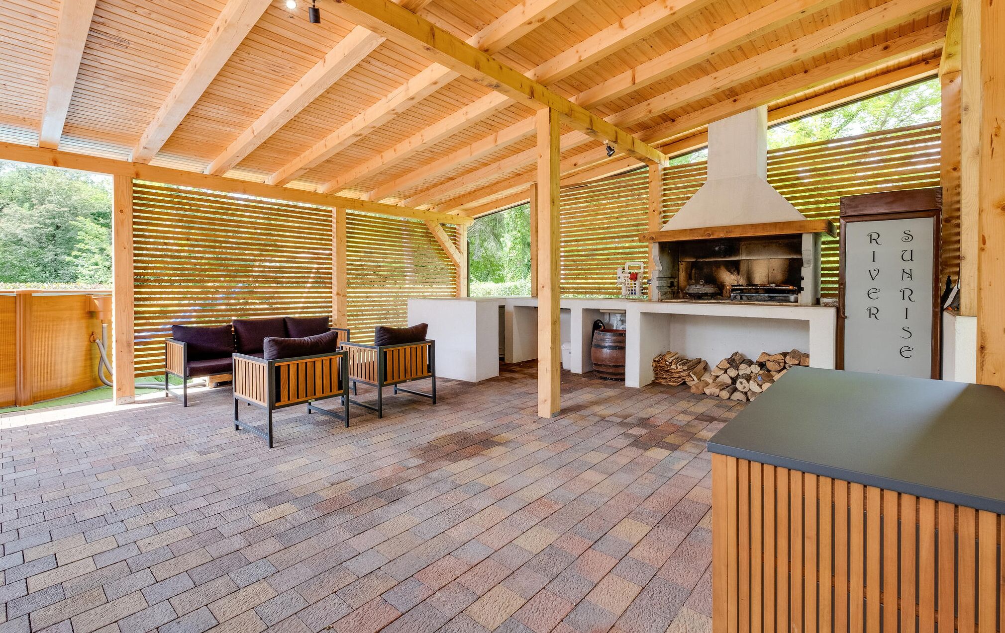 Un espace extérieur abrité doté d’un plafond en bois, d’un barbecue, de sièges avec des coussins et d’une cheminée.