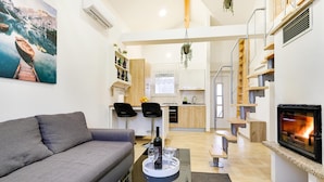 Een moderne woonkamer met een grijze bank, een salontafel met wijnglazen, een open haard en een keuken op de achtergrond.