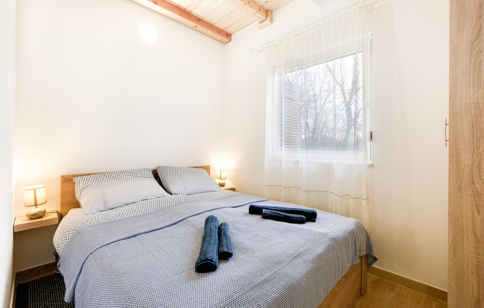 Ein ordentlich bezogenes Bett mit einem gemusterten Bettüberwurf und Kissen, eine Nachttischlampe und ein Fenster mit durchsichtigen Vorhängen.