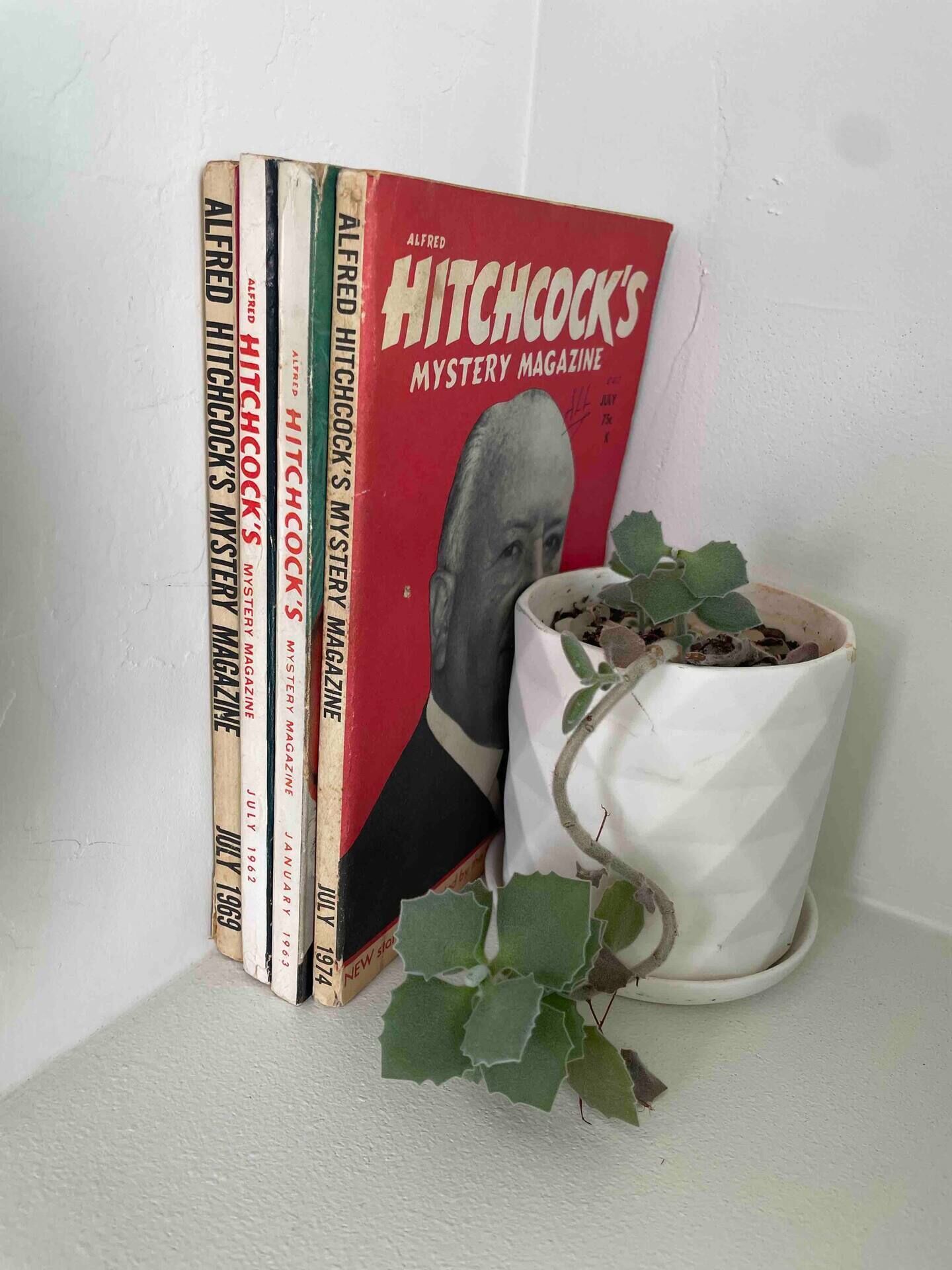 Une collection de livres intitulée « Hitchcock’s Mystery Magazine » et une plante en pot.