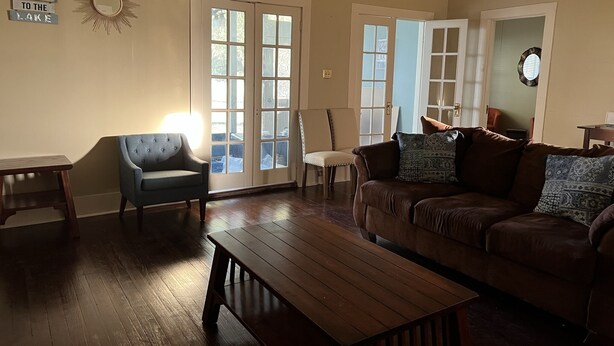 Sala de estar con una mesa de centro de madera, un sofá de cuero marrón, un sillón azul y una mesita auxiliar de madera. Hay dos puertas que dan a un balcón, y un espejo montado en la pared sobre la mesa de centro.