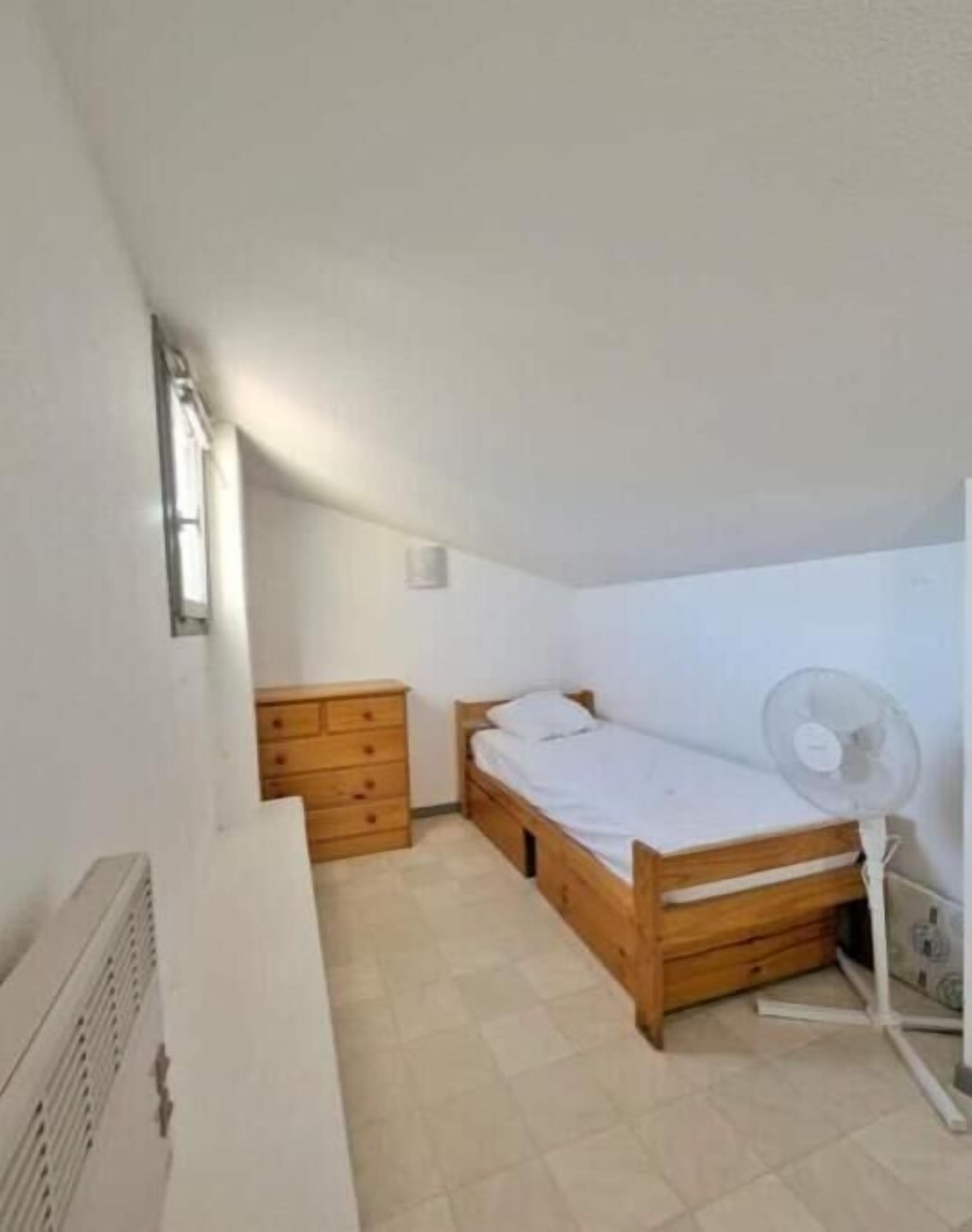 A small, single-room apartment with a wooden bed, a wooden dresser, a white radiator, and a standing fan.