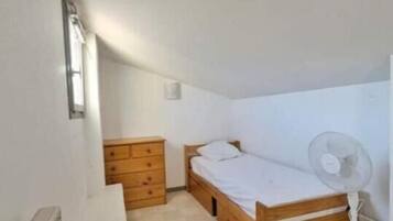 A small, single-room apartment with a wooden bed, a wooden dresser, a white radiator, and a standing fan.