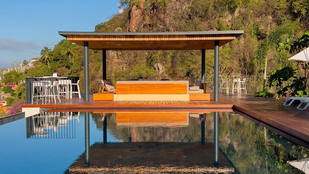 A infinity pool with a wooden deck and seating area, surrounded by lush greenery and a clear blue sky. - New Condo at Oasis 2 BD 2 Pools Riverside  2BD Condo for rent in El Caloso, Puer (Puerto Vallarta)