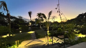 A well-lit outdoor area with a pathway leading to a building, surrounded by palm trees and greenery. - New Condo at Oasis 2 BD 2 Pools Riverside  2BD Condo for rent in El Caloso, Puer (Puerto Vallarta)