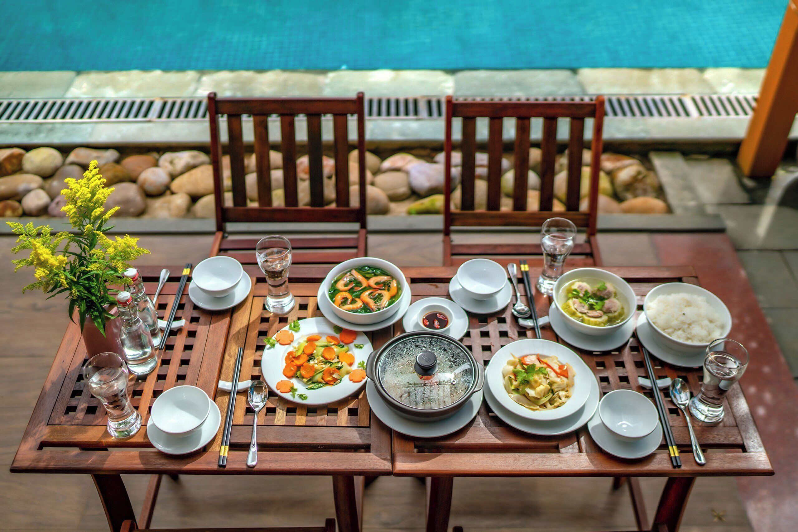 A dining table set with various dishes, utensils, and glasses, with a pool and rocks in the background.