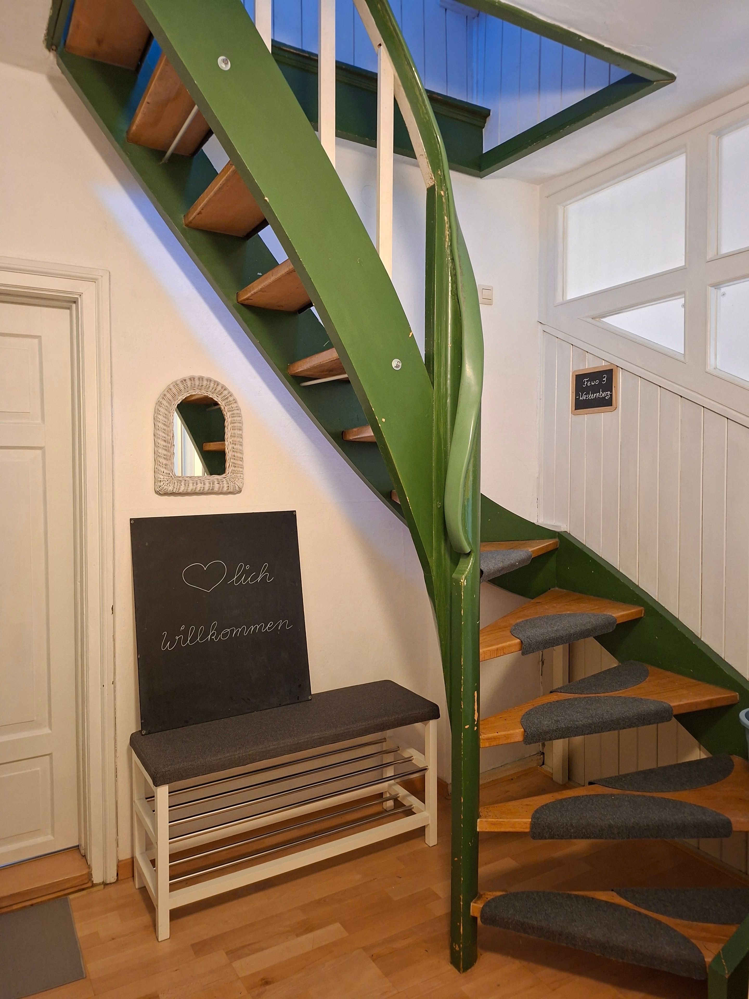 Eine Treppe mit grünen Geländern und Holzstufen, eine Tafel mit der Aufschrift 'welcome' und ein kleiner Spiegel an der Wand.