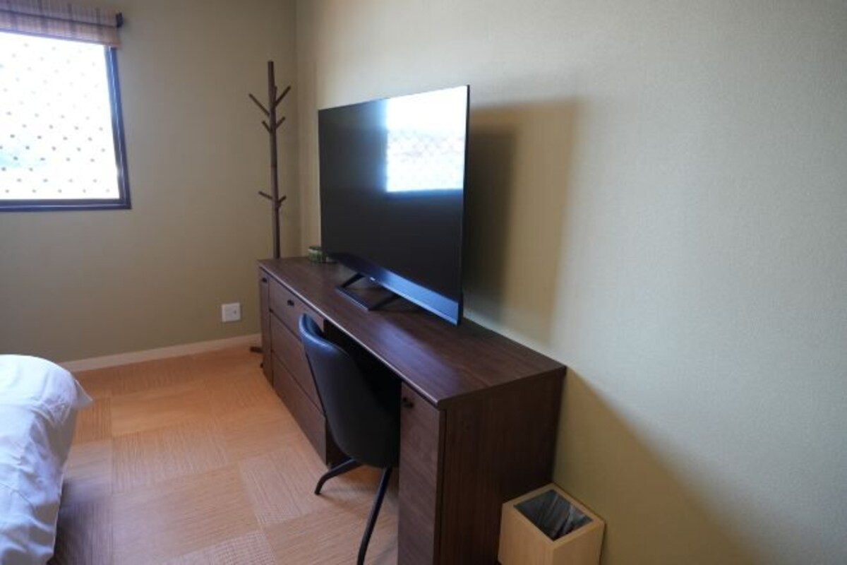 Un dormitorio con una cama, un escritorio de madera con un televisor de pantalla plana, una silla y un cubo de basura.