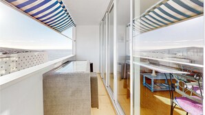 Un balcón con vista al mar, una mesa de vidrio, mobiliario de mimbre y toldos de tela a rayas.
