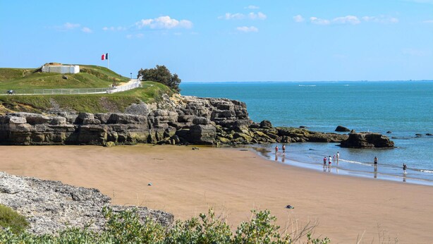 Paisaje costero con una playa arenosa, afloramientos rocosos y una colina cubierta de hierba con una estructura que muestra una bandera francesa.