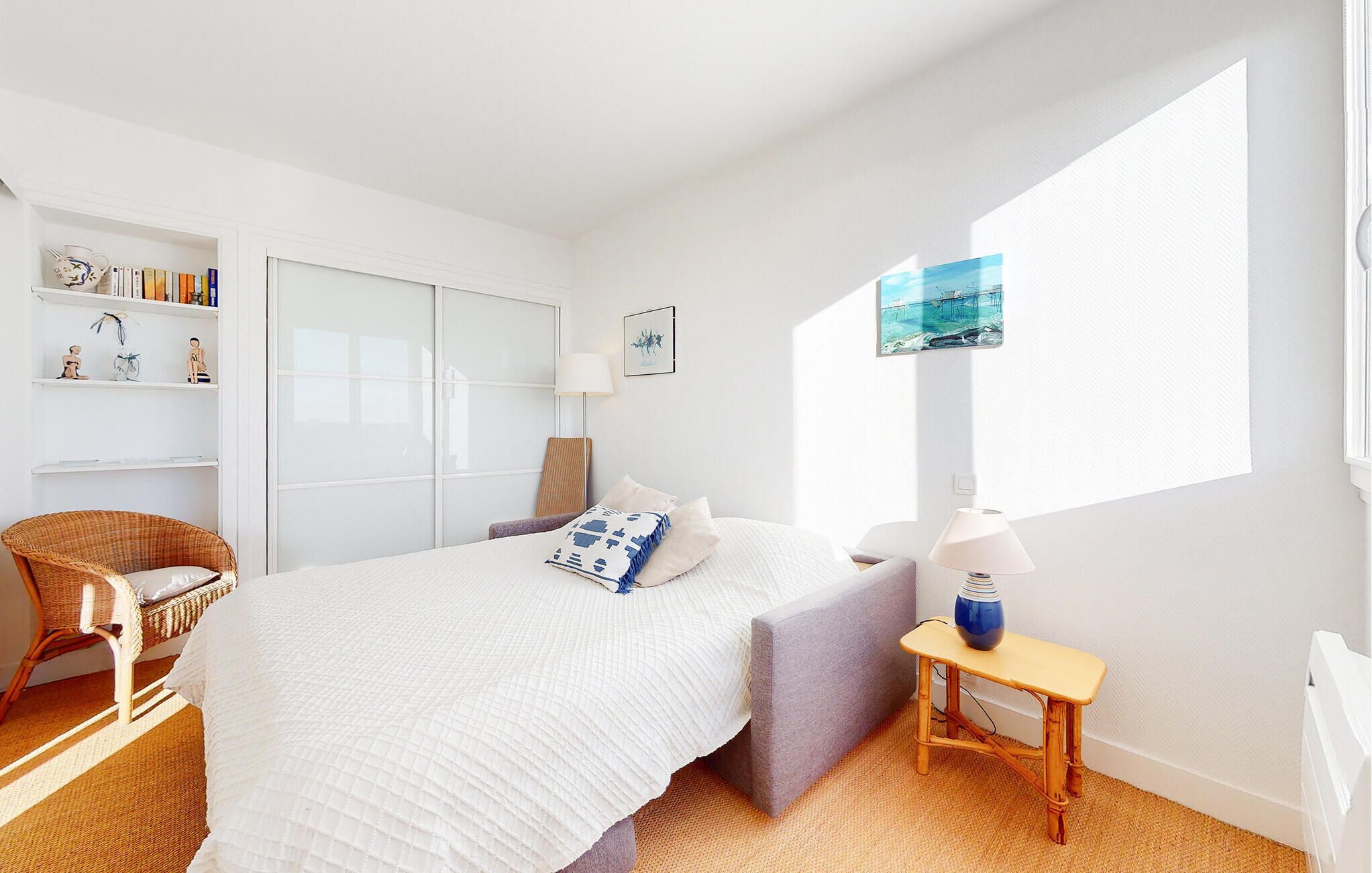 Un dormitorio con cama, una silla, una mesita de noche, una lámpara, una estantería con libros y objetos decorativos, y un ventanal con un cuadro.