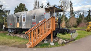 A stationary train caboose with a wooden staircase leading up to the door, situated on tracks with a grassy area and trees in the background.
