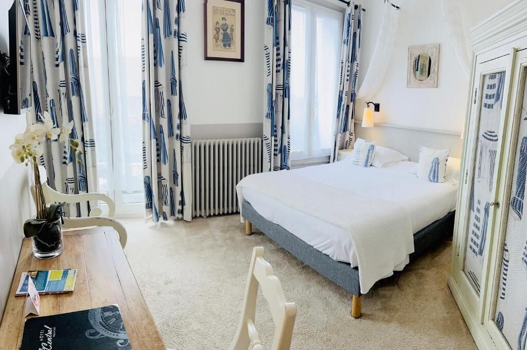 A bedroom with a bed, a radiator, a window with curtains, a desk with a chair, and a vase with flowers.