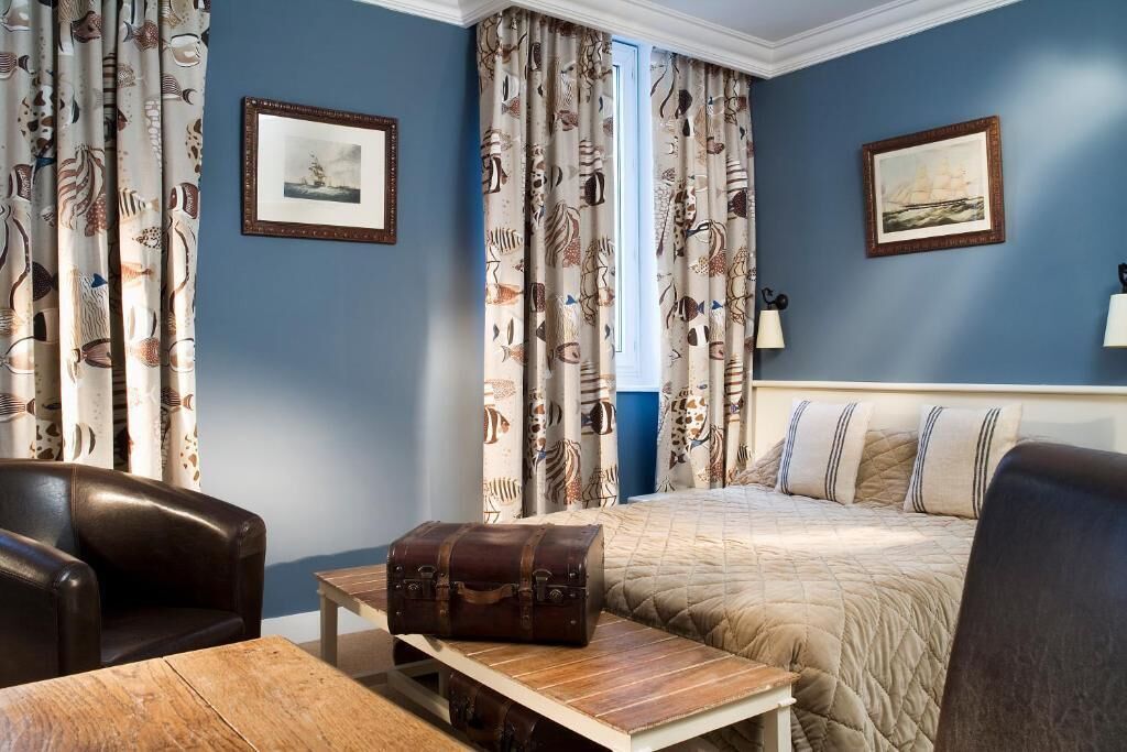 Habitación de hotel con cama, una mesa de madera, un sillón de cuero y cortinas con motivos náuticos.