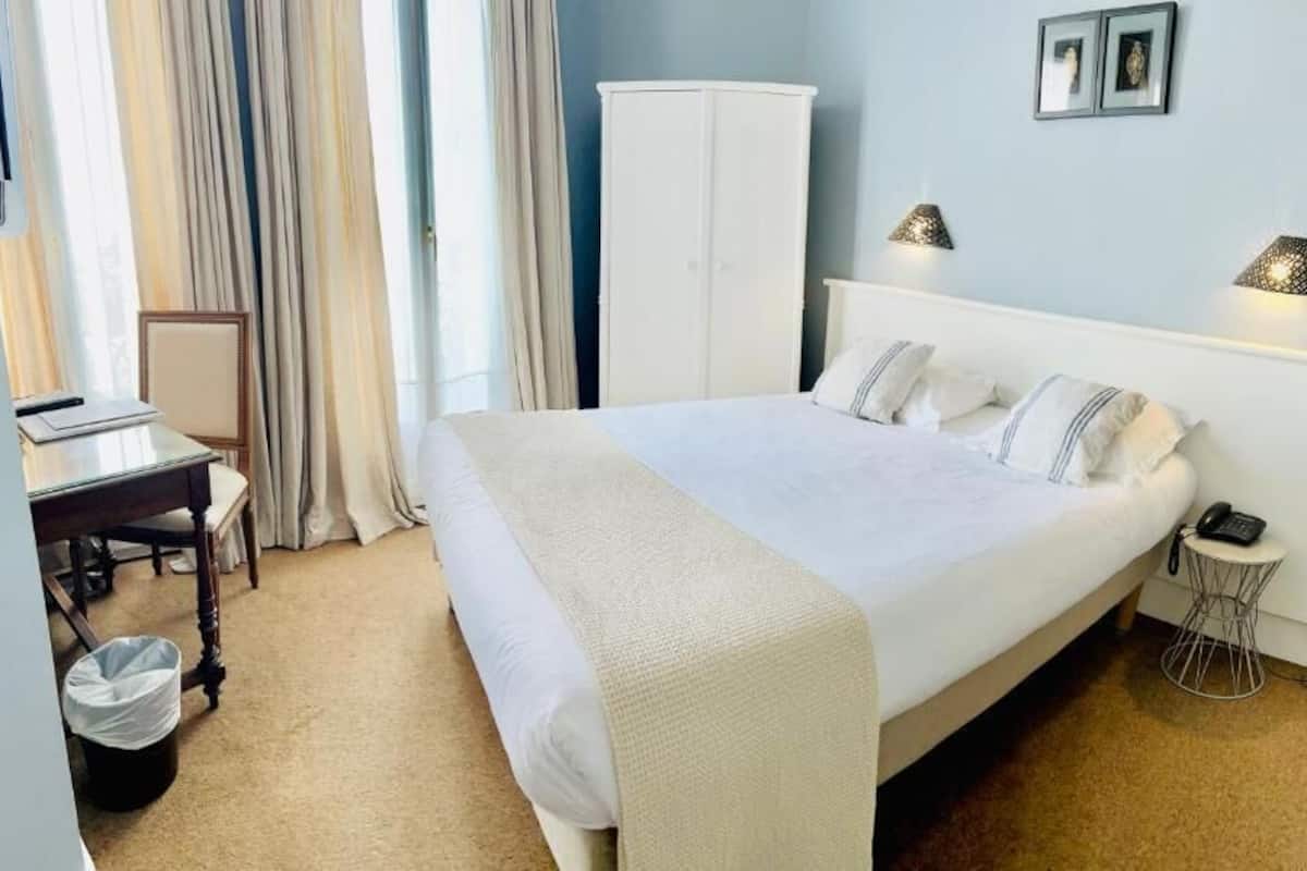 A hotel room with a bed, bedside table, lamp, wardrobe, and a desk with a chair.