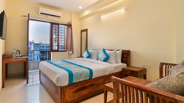 A bedroom with a wooden bed, a wooden desk, a chair, a television, and a balcony with a view of buildings. - Hotel The York (Noida)