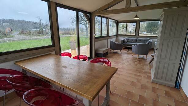 A spacious room with a large wooden table and red chairs, a grey sofa, and a view of the outdoors through large windows.