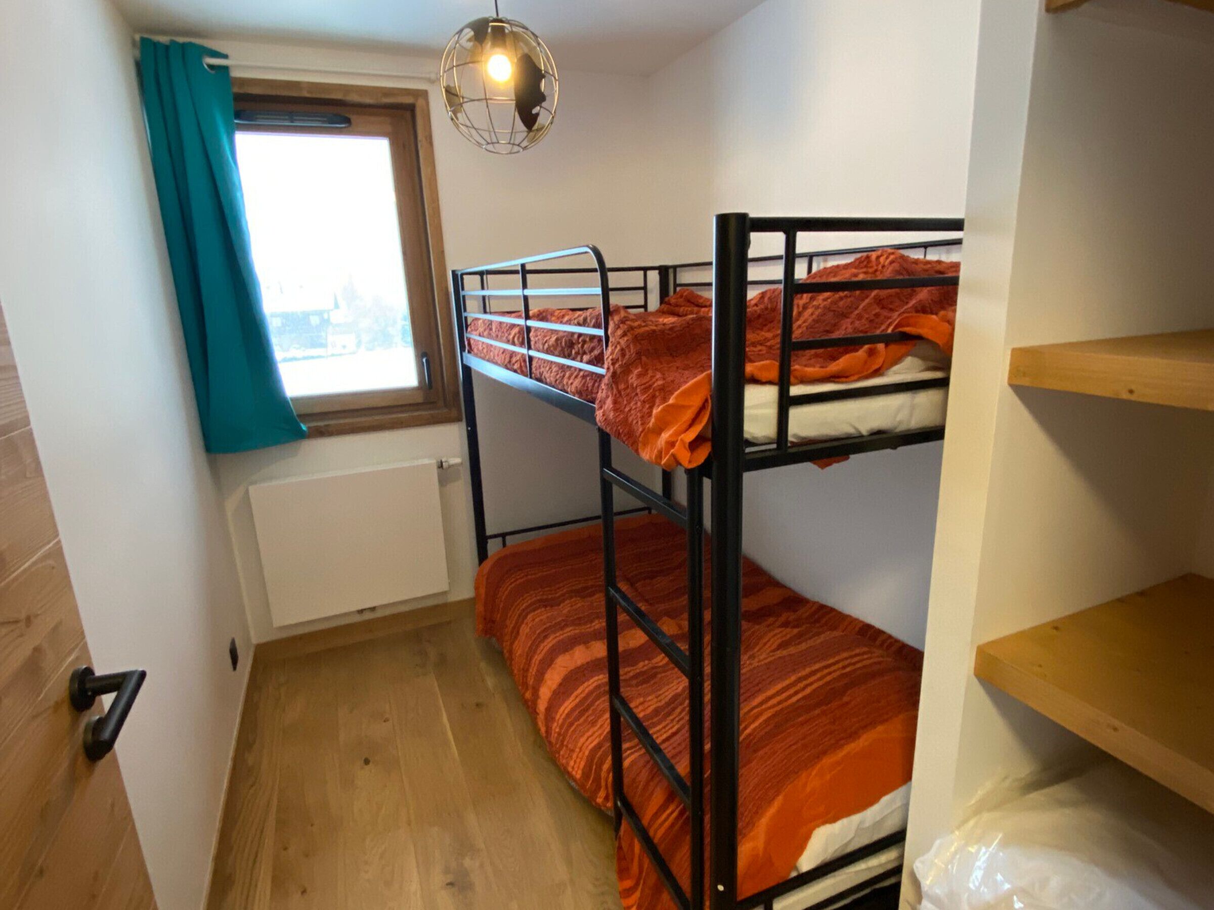 A small, compact room with bunk beds, a window with curtains, a radiator, and a wooden floor.