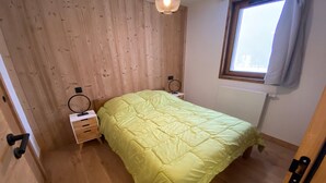 Une chambre avec un plancher en bois, un lit recouvert d’une courtepointe jaune, deux tables de chevet sur lesquelles sont posés des anneaux, une fenêtre avec des rideaux et un luminaire suspendu.