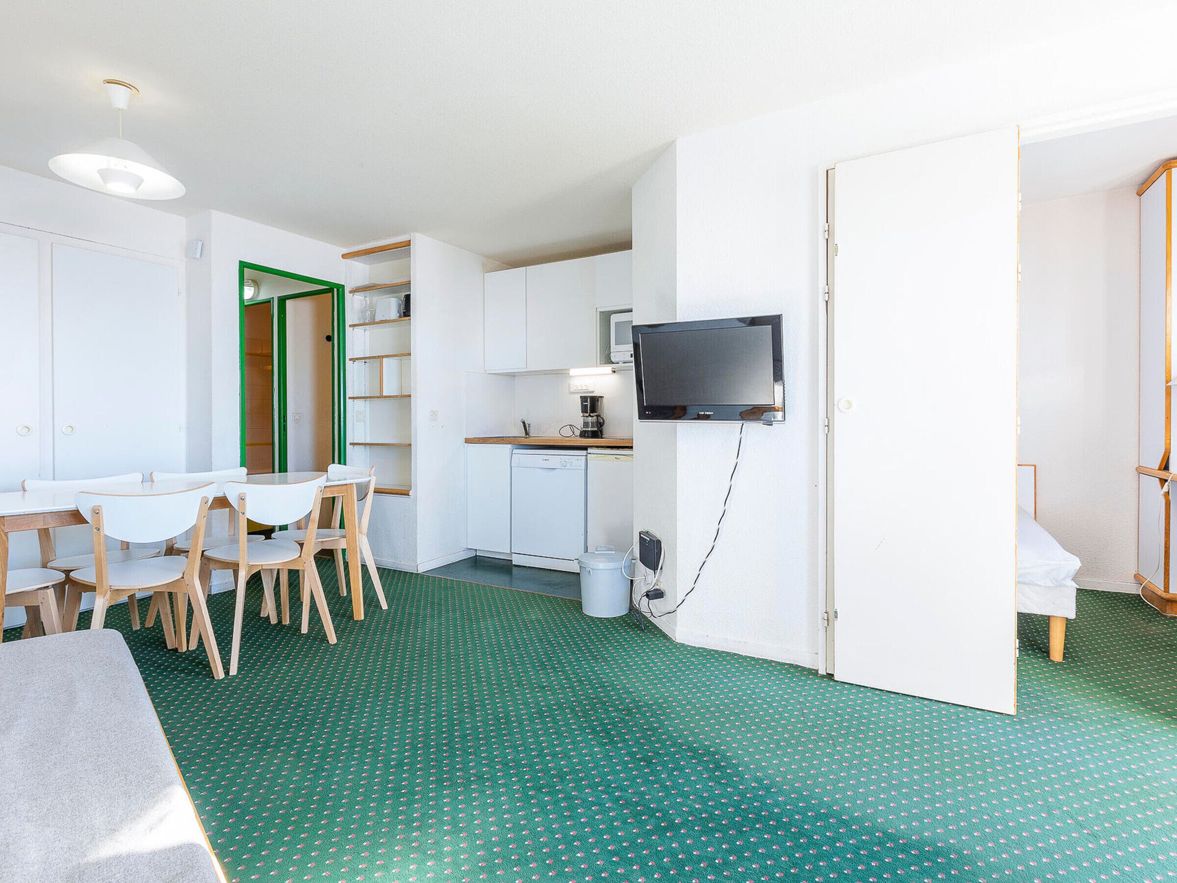 A compact room with a dining table, chairs, a small kitchenette, a television, and a green patterned floor.