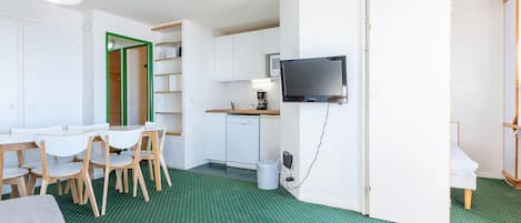 A compact room with a dining table, chairs, a small kitchenette, a television, and a green patterned floor.