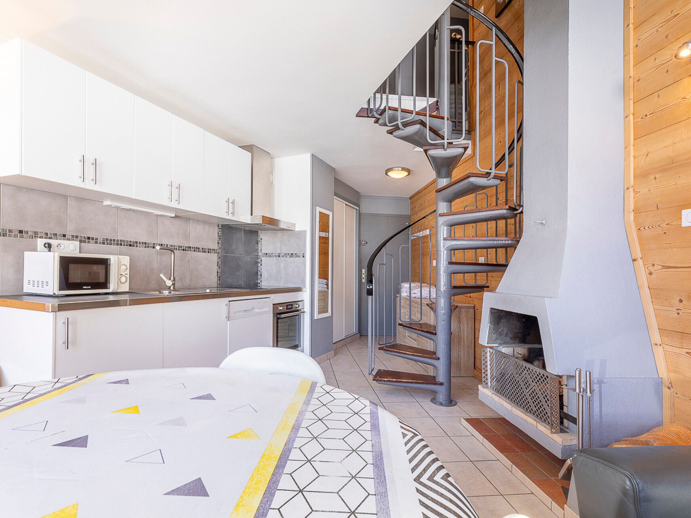 A modern kitchen with white cabinets, a microwave, and a built-in oven. There is a spiral staircase leading to a loft area with a fireplace.