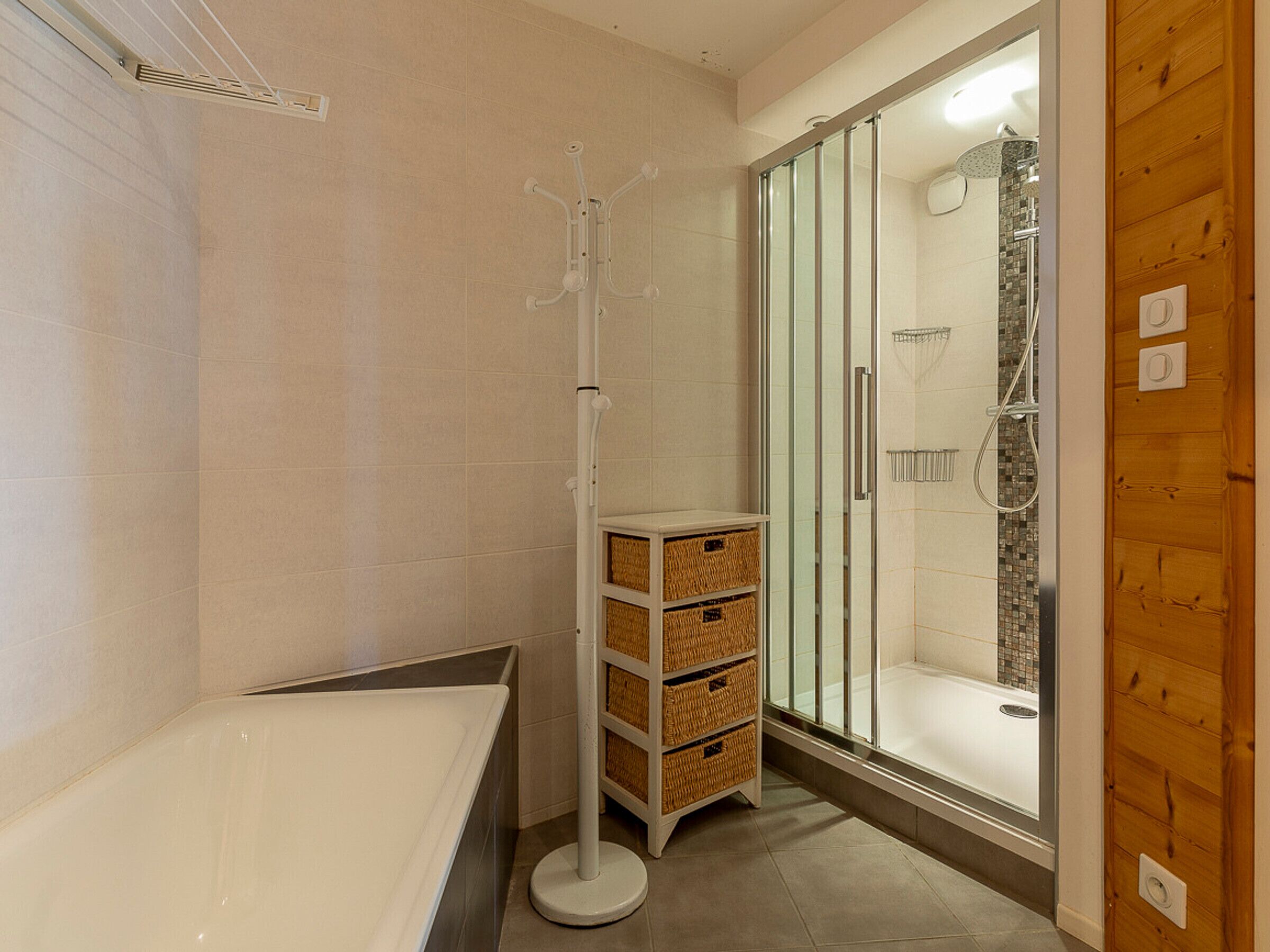 A modern bathroom with a glass-enclosed shower, a white bathtub, and wicker baskets on a white shelf.