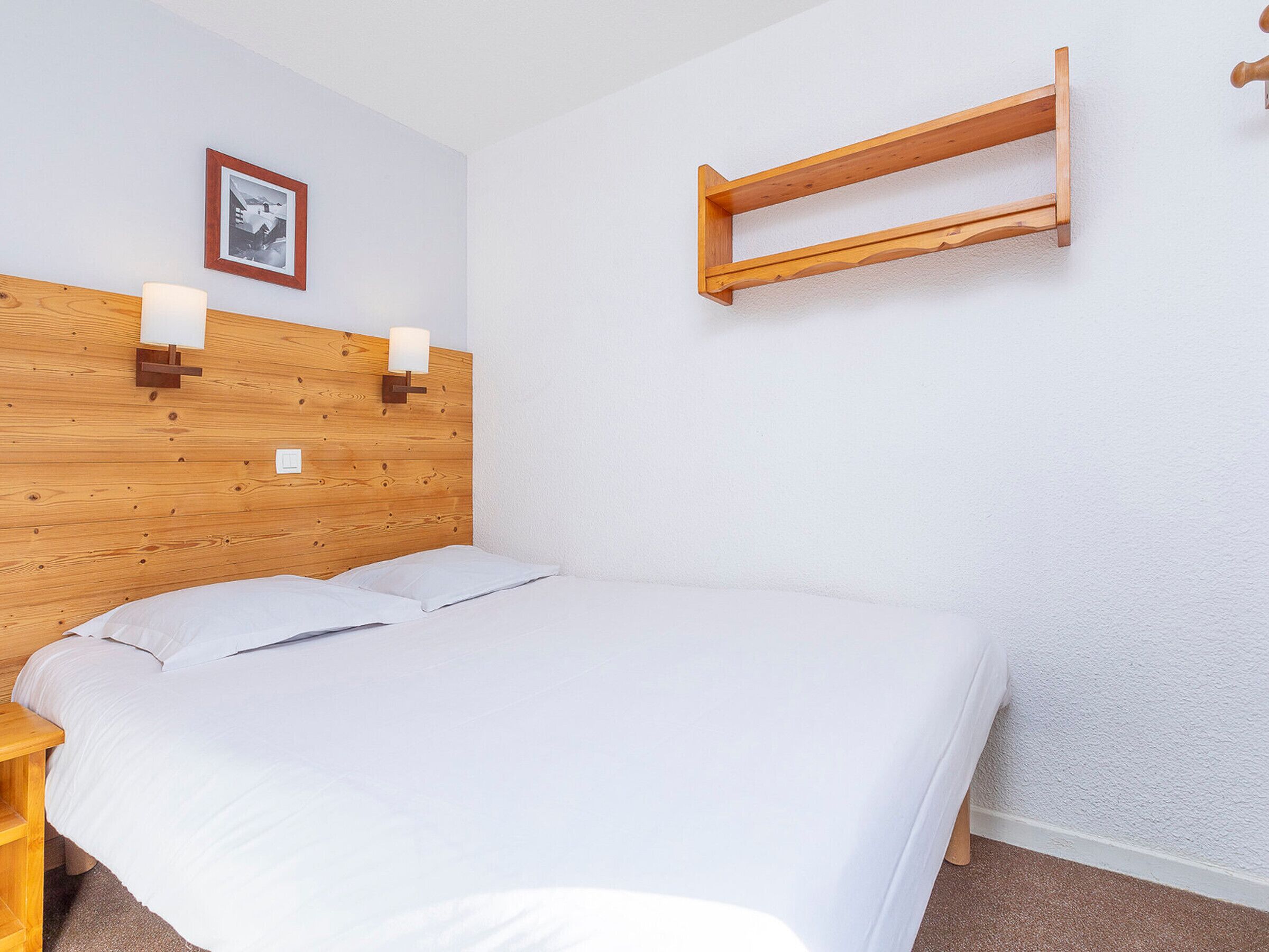A bedroom with a wooden headboard, two bedside lamps, a white bedspread, a wooden nightstand, and a small wooden shelf.
