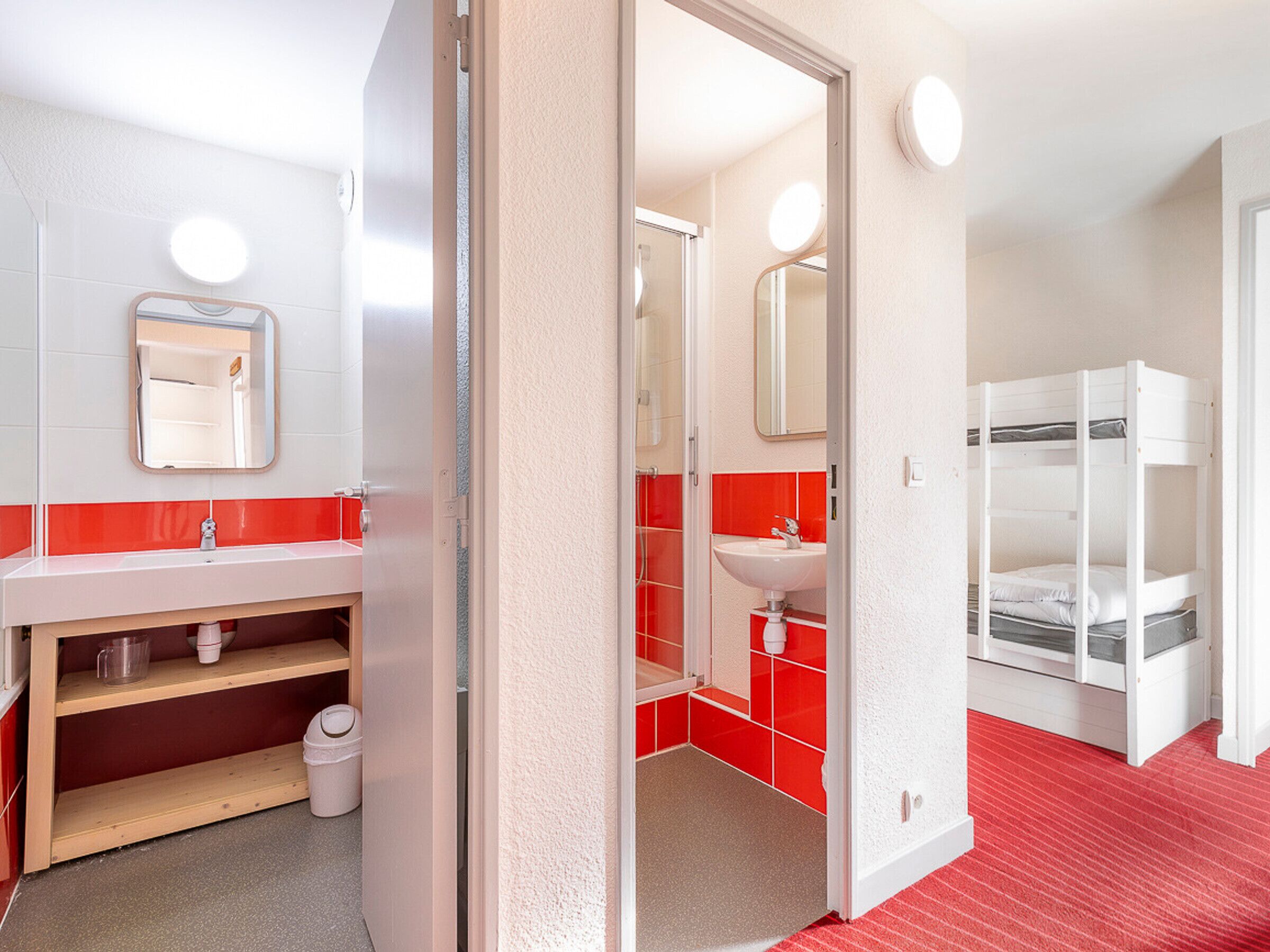 A hotel room with a red and white color scheme, including a red-tiled shower area, a white sink with a mirror, and a bunk bed with white bedding.