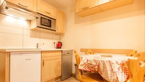 A compact kitchen with wooden cabinets, a microwave, a red kettle, a dishwasher, and a dining table with a checkered tablecloth.
