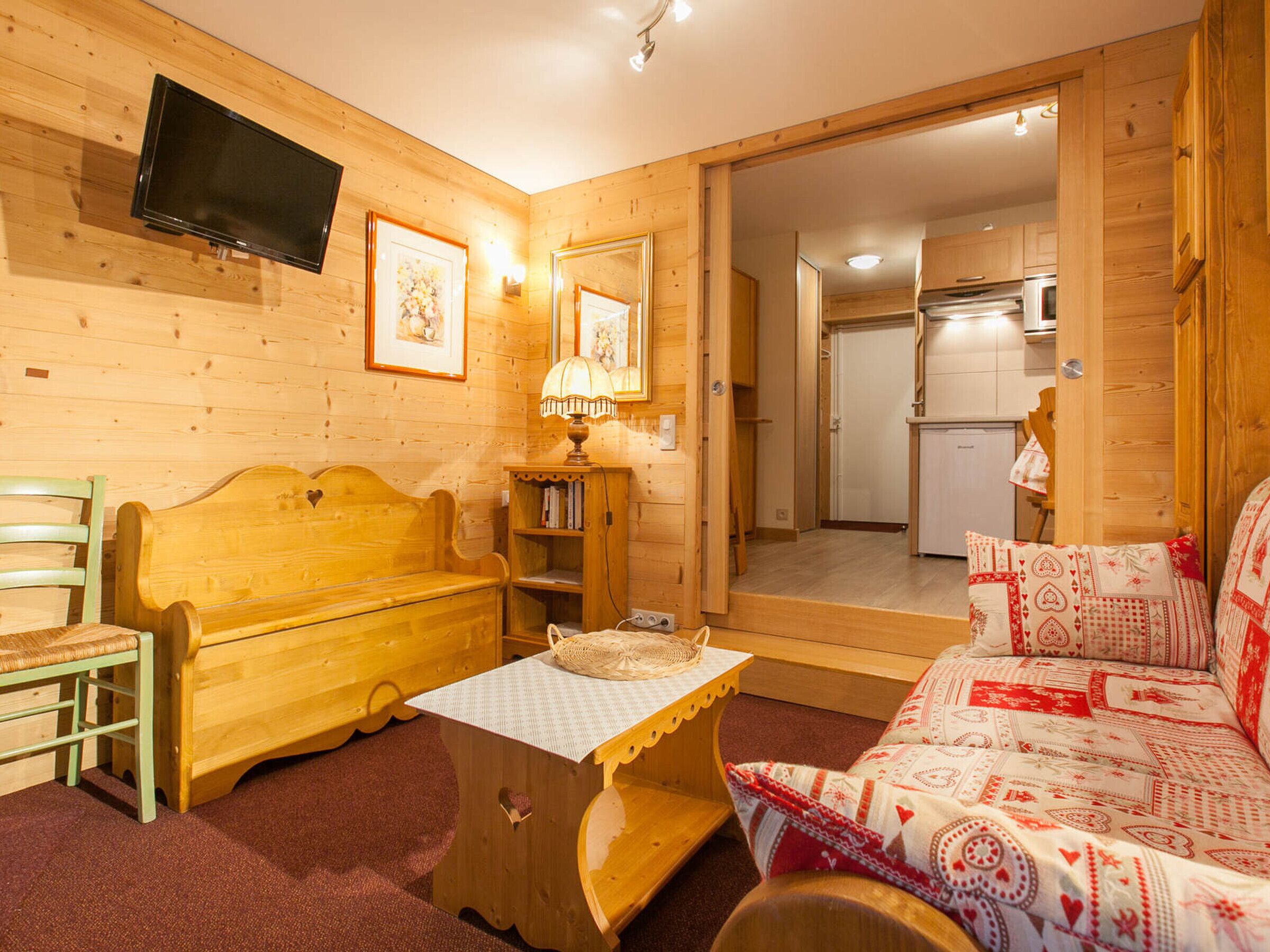 Une pièce chaleureuse avec un banc en bois, une petite table, un canapé agrémenté de coussins à motifs, un téléviseur fixé au mur et un coin cuisine visible à travers une porte ouverte.