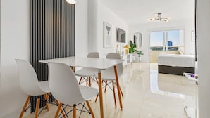 Ein moderner Essbereich mit einem weißen Tisch und Stühlen, einer vertikal gestreiften Wand und Blick auf den Wohnbereich mit Bett und Balkon.