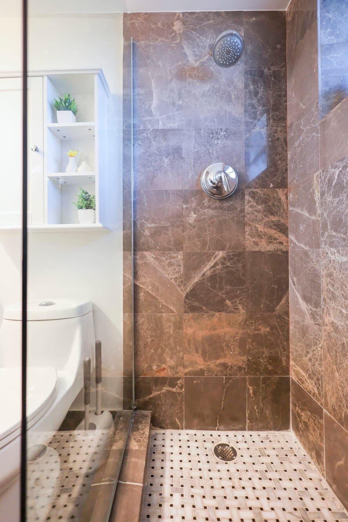 A bathroom with a glass-enclosed shower, marble tiles, a white sink, and a shelf with decorative items.