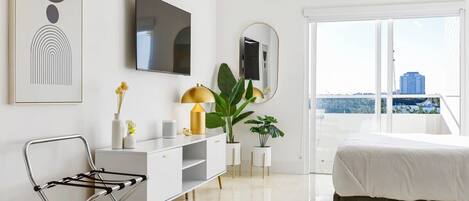 Een moderne slaapkamer met een bed, een witte ladekast, een televisie en een groot raam met uitzicht over de stad.