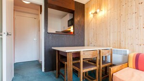 A compact room with a wooden table and chairs, a small kitchenette, and a cozy seating area with a cushion.