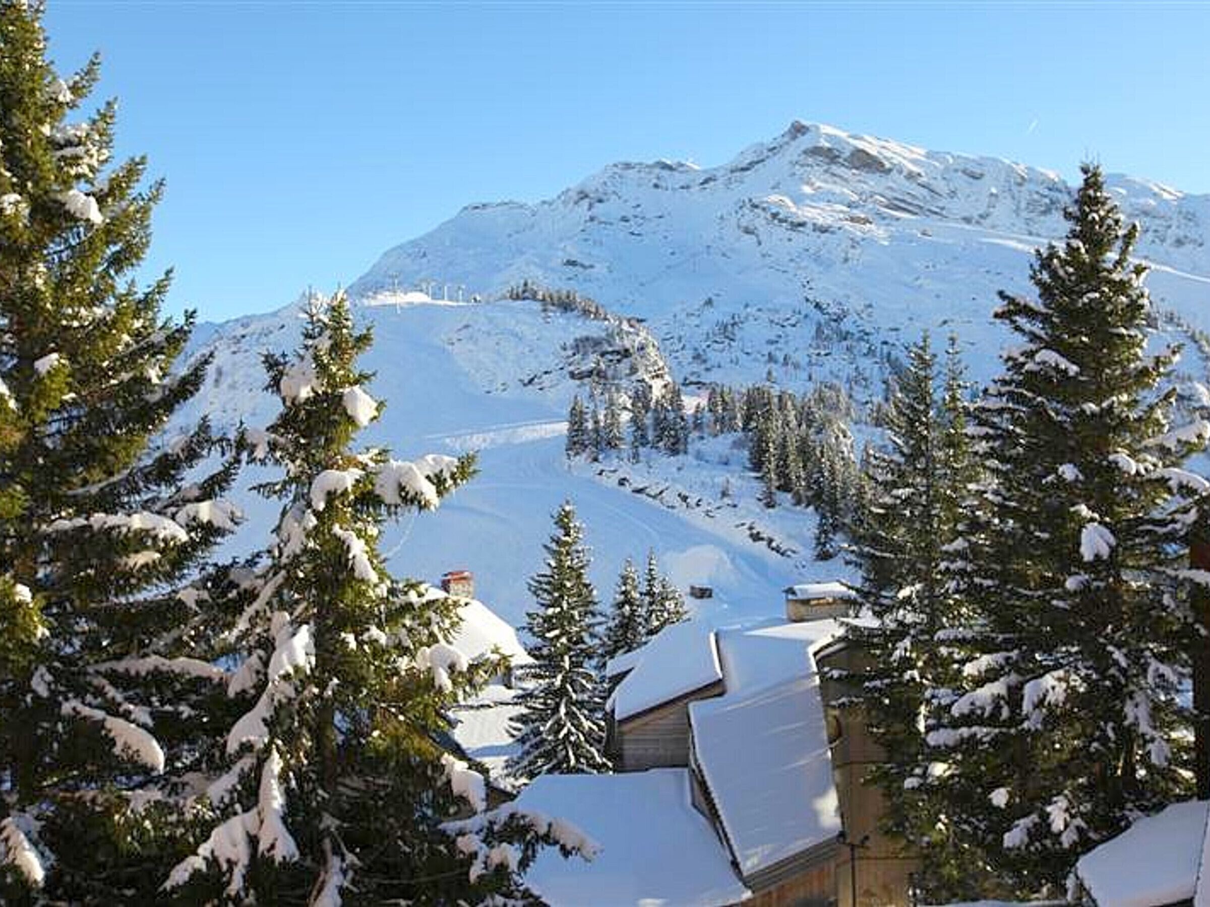 A snow-covered mountain with a clear sky. There are snow-covered trees and a building with a sloped roof.