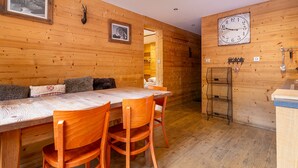 A wooden interior with a dining table and chairs, framed pictures, a wall clock, and a coat rack.