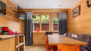 A cozy cabin interior with a wooden dining table, chairs, a sofa, and a television mounted on the wall.