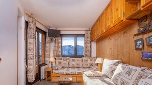 A cozy room with a wooden ceiling, a large window with a mountain view, a patterned sofa, and a wooden cabinet with pictures.