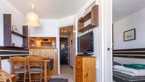 Une pièce de taille modeste, avec une table et des chaises en bois, un téléviseur posé sur un meuble en bois, un lit recouvert d’une couverture verte et un petit coin cuisine doté d’un comptoir en bois et d’un luminaire suspendu.