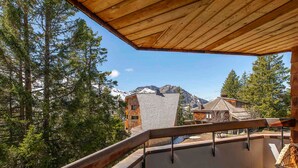 Een houten balkon met metalen hekwerk, met uitzicht op een bergachtig landschap met bomen en gebouwen.